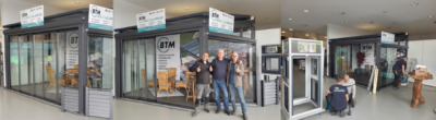 Ein Foto des BTM Holz + Aluminium-Teams bei der Modernisierungsmesse in Osterholz-Scharmbeck. Drei Teammitglieder, dieselben wie in image_3.png, stehen vor ihrem Messestand-Modell. Ein Mitglied erklärt dem anderen Details der Aluminium-Struktur, während das dritte Mitglied daneben steht und auf einen kleinen Banner mit den Dienstleistungssymbolen (Terrasse, Wintergarten, Carport) zeigt. Der obere Banner des Standes zeigt das BTM-Logo und den Namen des Unternehmens: BTM HOLZ + ALUMINIUM, mit der Telefonnummer (04791-5024 449). Im Hintergrund sind die Hallenumgebung und andere Ausstellungsstände zu sehen. Die Teammitglieder lächeln und interagieren.
