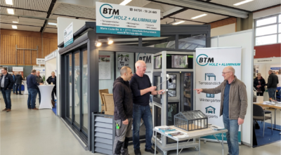 Das Team von BTM Holz + Aluminium steht gut gelaunt mit "Daumen hoch" vor dem Messestand in der Stadthalle Osterholz-Scharmbeck. Im Hintergrund ist ein modernes Terrassendach-Modell sowie das Firmenlogo mit Informationen zu Wintergärten und Carports zu sehen. Die Teammitglieder tragen dunkle Arbeitskleidung und Jeans.